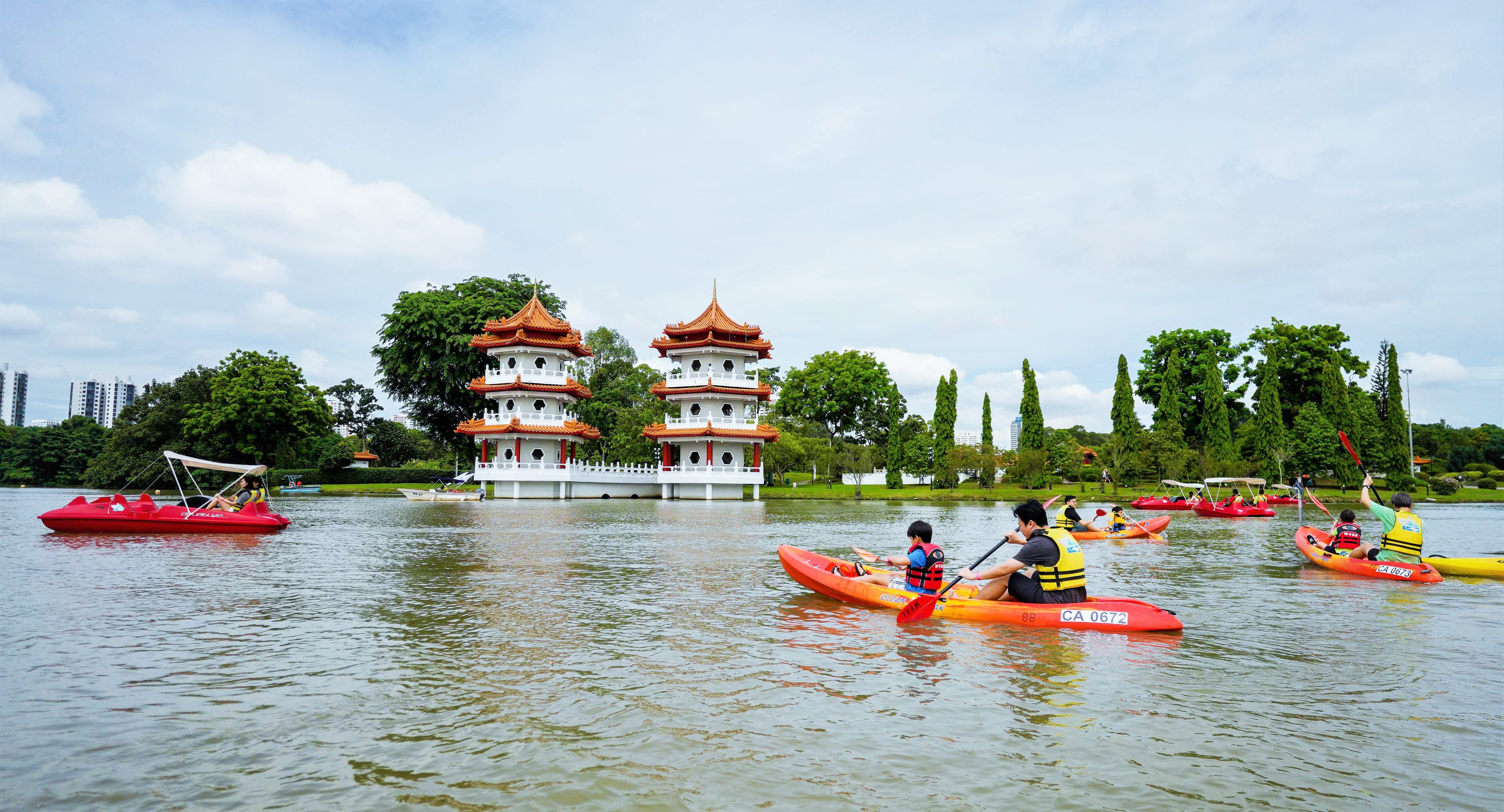 People are kayaking on a lake with traditional pagodas and lush trees in the background under a partly cloudy sky.